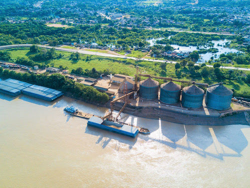 Logística amazônica avança com práticas sustentáveis e gestão ambiental integrada A logística portuária na Amazônia tem avançado na adoção de práticas sustentáveis que conciliam eficiência operacional, controle ambiental e desenvolvimento social. Por meio de ações integradas e de uma governança ambiental compartilhada, os associados da Associação dos Terminais Portuários e Estações de Transbordo de Carga da Bacia Amazônica, AMPORT, vêm fortalecendo um modelo de atuação que reduz impactos ambientais e amplia os benefícios para as comunidades do entorno. Por meio do Plano de Controle Ambiental Integrado (PCAI), plano que orienta ações de controle, monitoramento e mitigação dos impactos associados às atividades portuárias, descarga, armazenamento e transbordo de grãos na região de Miritituba (PA), os associados garantem o atendimento às condicionantes do licenciamento ambiental, a harmonização das ações entre os empreendimentos e uma gestão ambiental mais integrada na região. De acordo com Caroline Arruda, coordenadora de consultoria do PCAI, as práticas sustentáveis adotadas pelas empresas associadas abrangem desde a gestão de resíduos e efluentes até o monitoramento contínuo da qualidade do ar, das águas superficiais, do ruído ambiental e da fauna terrestre e aquática. “A implementação do PCAI trouxe avanços técnicos relevantes, como a padronização dos procedimentos de monitoramento ambiental, o aprimoramento dos mecanismos de controle e mitigação de impactos, a maior integração entre os associados e o fortalecimento da governança ambiental”, explica. Entre os indicadores priorizados estão o controle de emissões atmosféricas difusas, especialmente de material particulado, o monitoramento dos recursos hídricos, com foco no Rio Tapajós e no Igarapé Santo Antônio, e o acompanhamento da biodiversidade. No campo social, o PCAI estrutura ações de comunicação social, educação ambiental e relacionamento institucional, garantindo diálogo permanente com comunidades do entorno e atenção às demandas da comunidade pesqueira. Além da gestão ambiental integrada, a adoção do modal hidroviário é apontada como uma das principais práticas sustentáveis da logística amazônica. Segundo o diretor-presidente da AMPORT, Flávio Acatauassú, o uso predominante das hidrovias torna a cadeia logística mais eficiente do ponto de vista energético e ambiental. “Essa adoção, por si só, já representa a redução de emissões e a maior eficiência energética, além de mitigar os impactos gerados por outros modais de transporte”, afirma. Os resultados desse modelo já podem ser mensurados. A substituição de trechos rodoviários pelo transporte hidroviário nos corredores Branco–Negro, Guamá–Capim, Tocantins e Tapajós resultou em reduções de emissões de gases poluentes de 90,87%, 70,28%, 34,83% e 22,09%, respectivamente. Para os próximos anos, a AMPORT projeta avanços concentrados no aprimoramento contínuo da gestão integrada do PCAI, na qualificação e ampliação dos indicadores socioambientais, no fortalecimento dos programas sociais e no aprofundamento das ações de monitoramento ambiental. “Já consolidamos um modelo mais sustentável com o modal fluvial. O desafio agora é garantir a perenidade da navegação, inclusive nos períodos de seca, e complementar a malha existente com a implantação de ferrovias”, afirma o diretor-presidente. Ao integrar eficiência logística, controle ambiental, inovação e desenvolvimento social, a atuação dos associados da AMPORT reforça o papel da logística amazônica como um vetor estratégico para o desenvolvimento sustentável da região, com soluções concretas que conciliam competitividade e responsabilidade socioambiental.